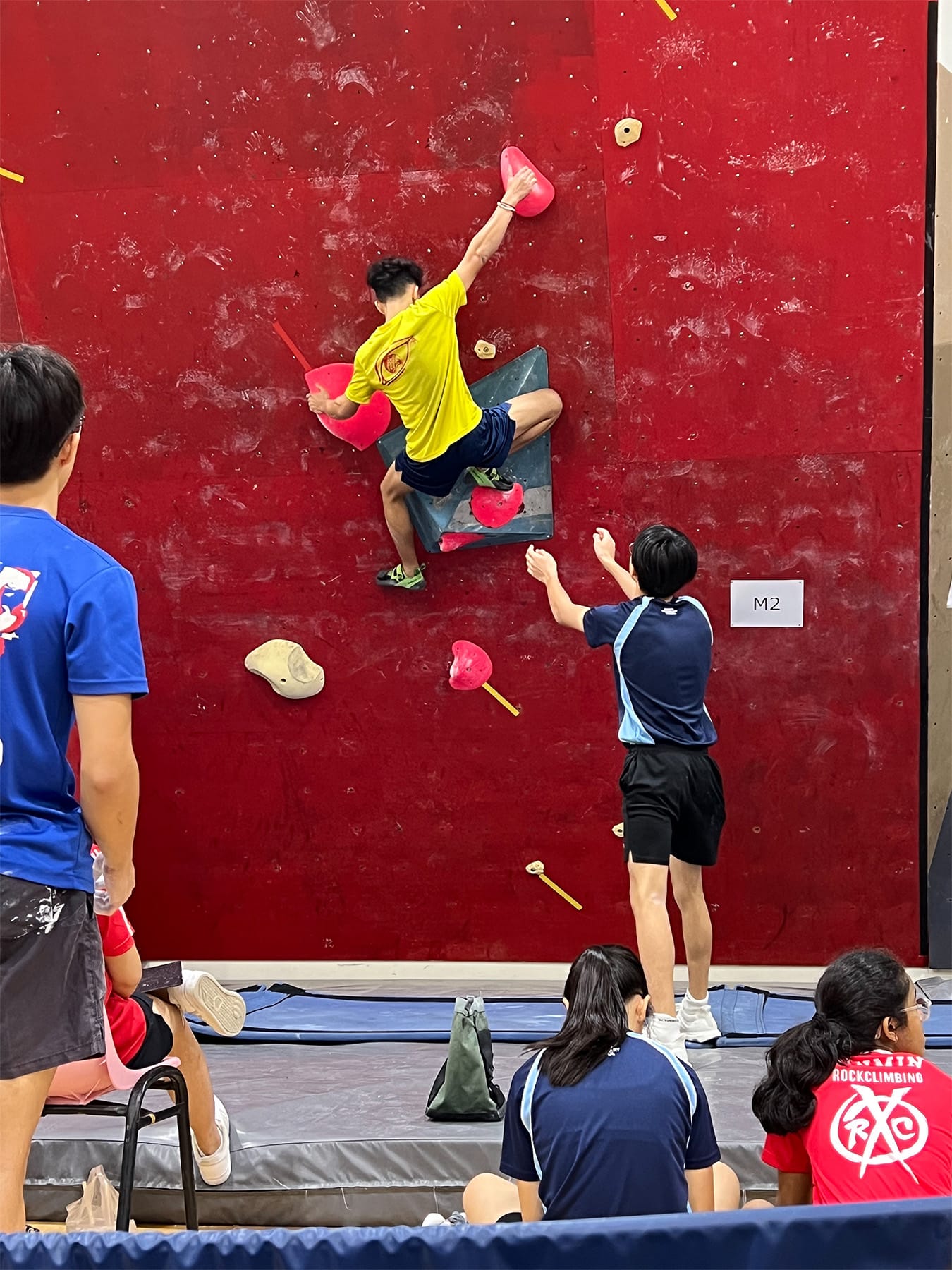 A youth in a yellow shirt climbs a red bouldering wall with pink holds as spectators watch below.